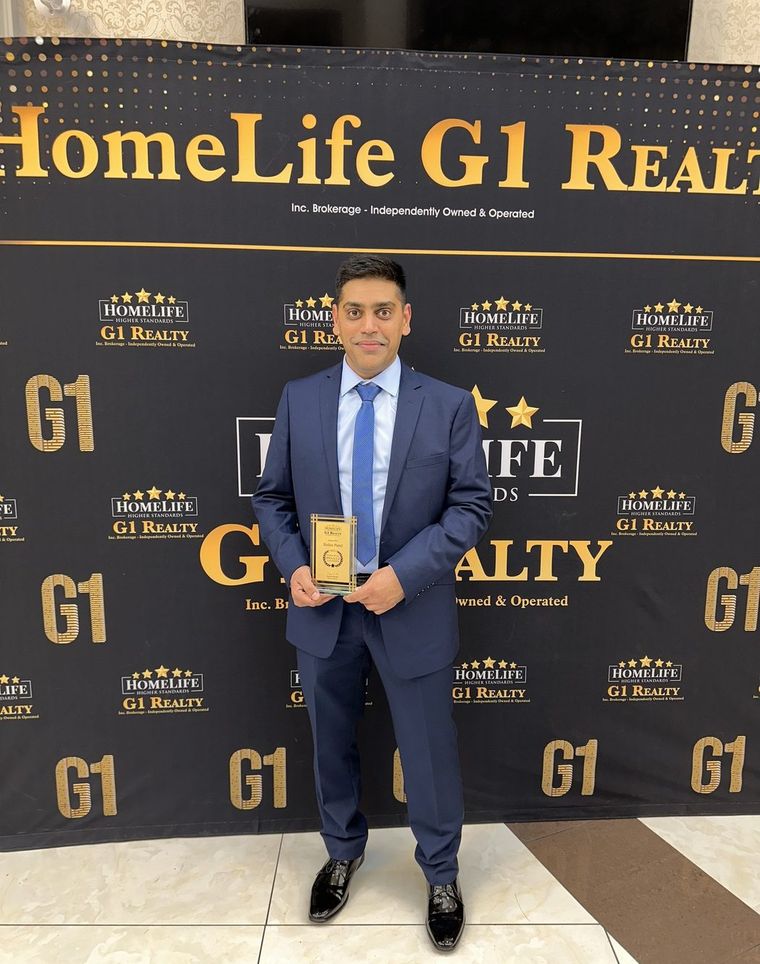 Robin Patel in a navy suit holding an award plaque in front of a HomeLife G1 Realty backdrop.