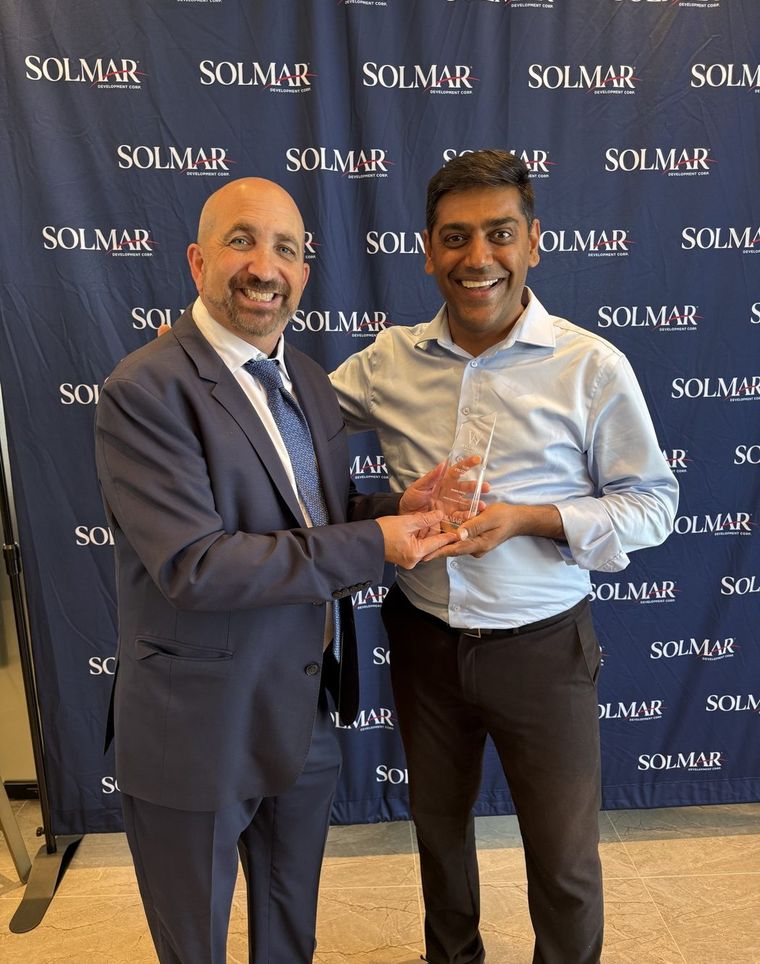Robin Patel shaking hands and holding a glass award in front of a Solmar backdrop.